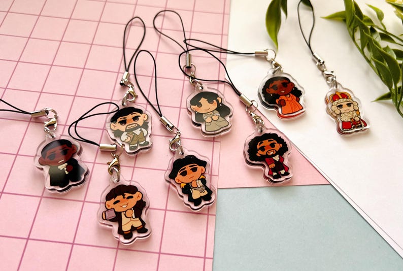 K&ouml;nnte beinhalten: Acht bunte Comicfiguren-Anh&auml;nger an schwarzen Schnur-Handy-Anh&auml;ngern. Die Figuren stammen aus dem Musical Hamilton.