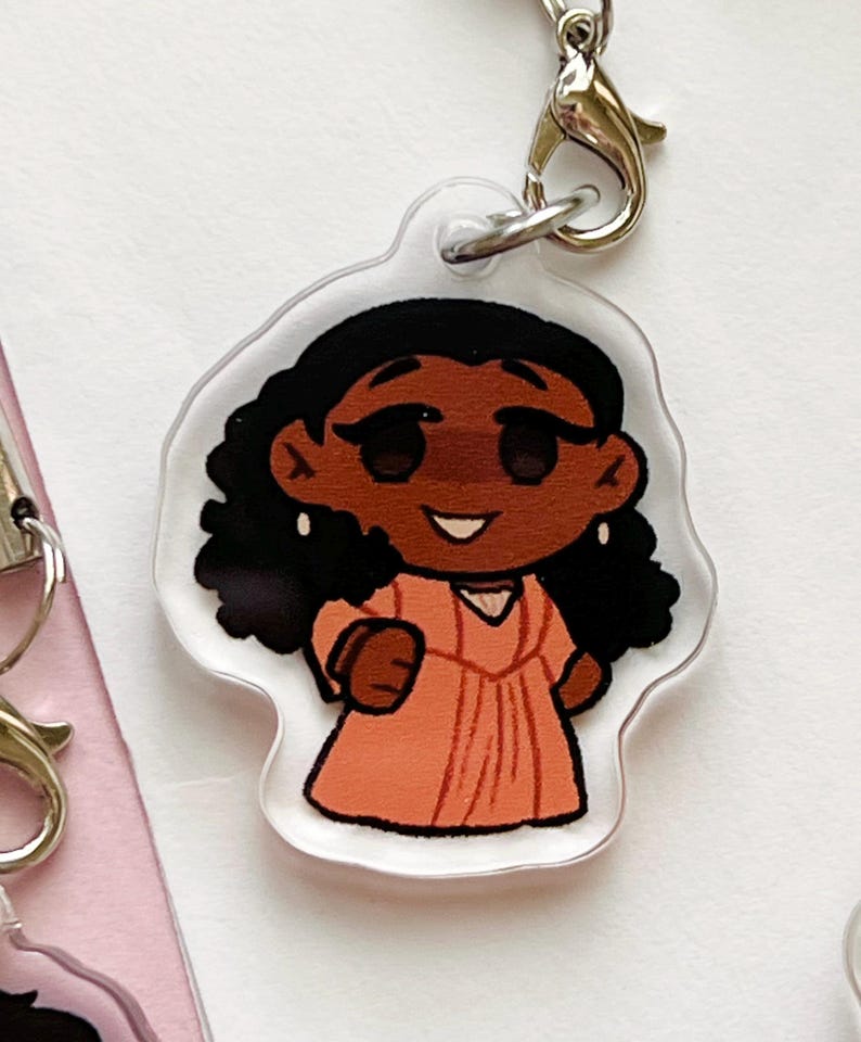 K&ouml;nnte beinhalten: Ein durchsichtiger Acryl-Schl&uuml;sselanh&auml;nger mit einer Cartoon-Illustration einer Frau mit brauner Haut, dunklen lockigen Haaren und einem rosa Kleid. Sie l&auml;chelt und hat gro&szlig;e Augen.