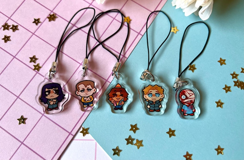 May include: Five clear acrylic charms with black string attached. Each charm features a different cartoon character. The characters are a girl with purple hair, a boy with brown hair, a girl with red hair, a boy with blonde hair, and a boy with a bandage on his head.