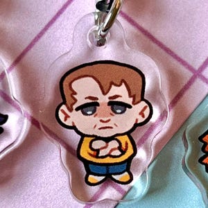 May include: A cartoon character keychain with a brown hair, yellow shirt, blue jeans, and white shoes. The character has a serious expression on their face.