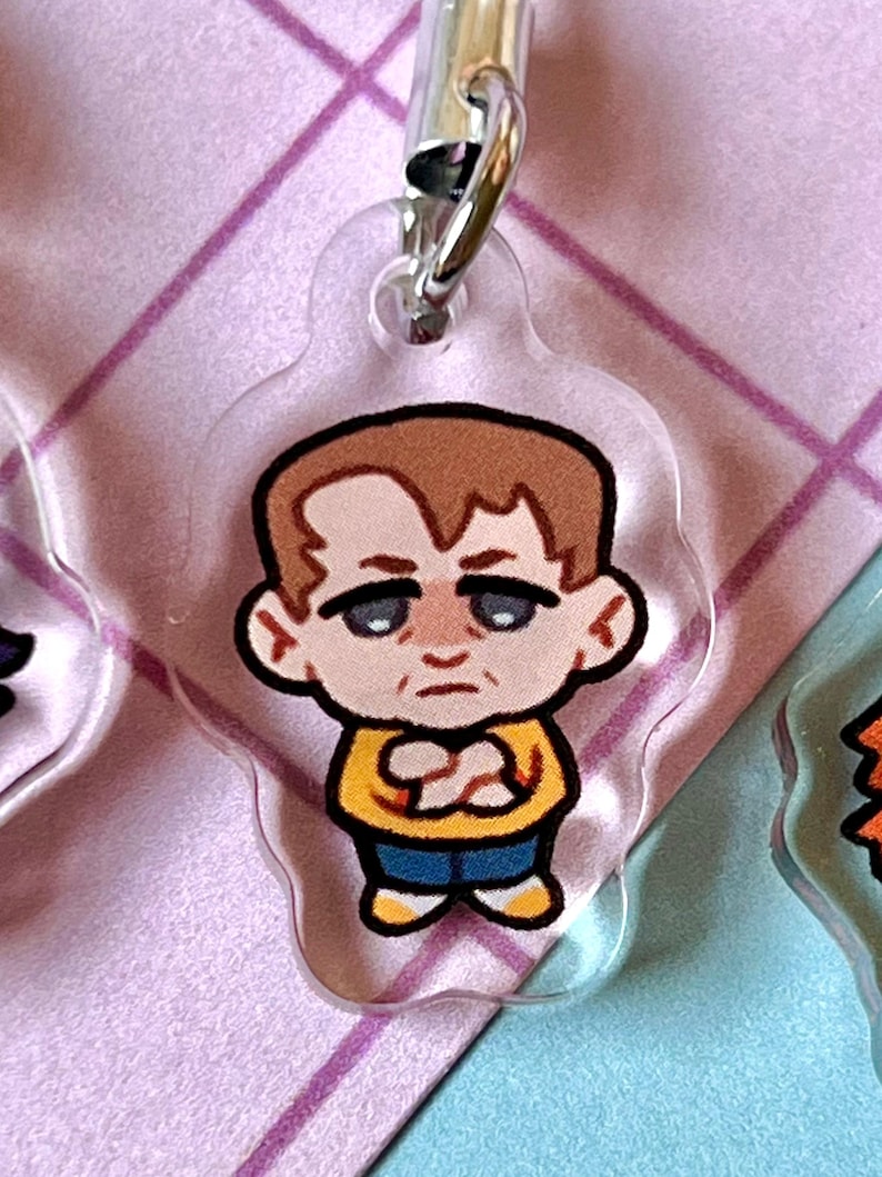 May include: A cartoon character keychain with a brown hair, yellow shirt, blue jeans, and white shoes. The character has a serious expression on their face.