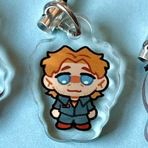 May include: A clear acrylic keychain with a cartoon illustration of a person with blond hair, blue eyes, and a red and blue jacket. The person is wearing a blue jumpsuit and red shoes.