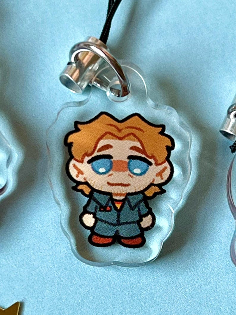 May include: A clear acrylic keychain with a cartoon illustration of a person with blond hair, blue eyes, and a red and blue jacket. The person is wearing a blue jumpsuit and red shoes.
