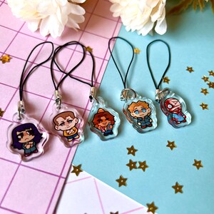 May include: Five colorful cartoon character charms on black string phone charms. The charms are clear plastic with printed images of people with different hairstyles and clothing.
