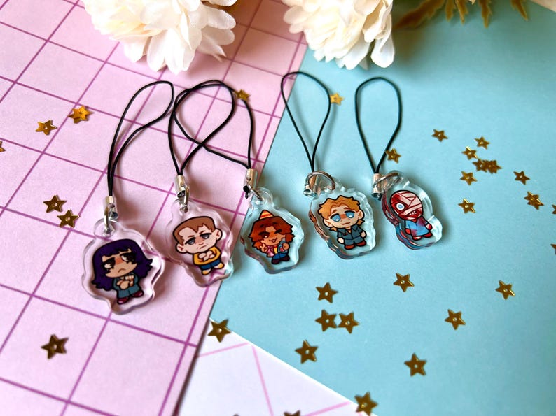 May include: Five colorful cartoon character charms on black string phone charms. The charms are clear plastic with printed images of people with different hairstyles and clothing.