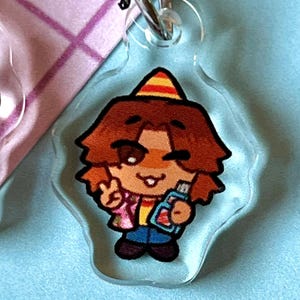 May include: A clear acrylic keychain featuring a cartoon character wearing a party hat, a pink and yellow shirt, blue jeans, and holding a blue and red object. The character has brown hair and is smiling.