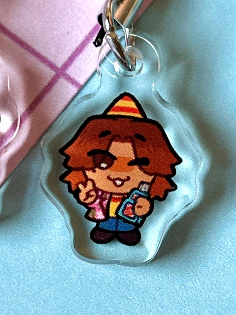 May include: A clear acrylic keychain featuring a cartoon character wearing a party hat, a pink and yellow shirt, blue jeans, and holding a blue and red object. The character has brown hair and is smiling.