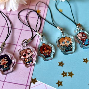 May include: Five colorful character charms on black string phone charms. The characters are drawn in a cartoon style and are wearing different outfits. The charms are all different shapes and sizes.