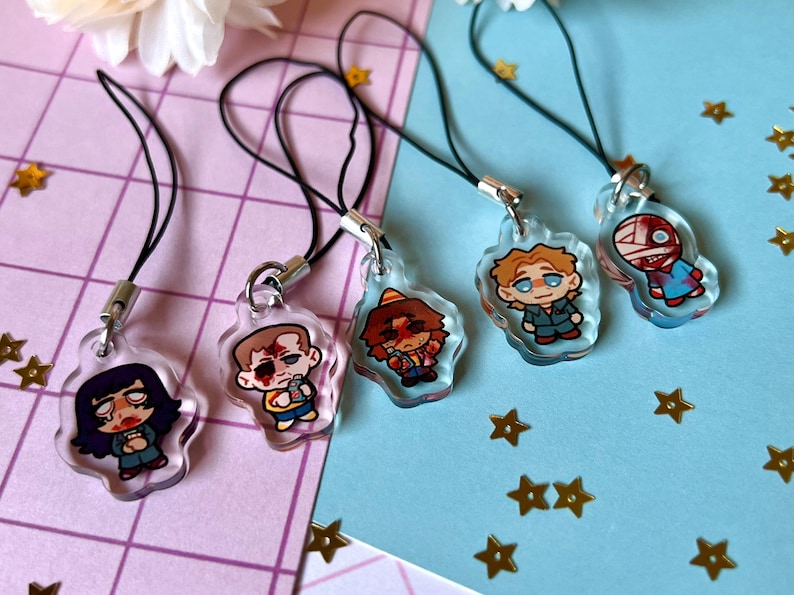 May include: Five colorful character charms on black string phone charms. The characters are drawn in a cartoon style and are wearing different outfits. The charms are all different shapes and sizes.