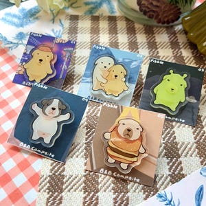 May include: A collection of five clear acrylic pins featuring cartoon dog illustrations. Each pin has a unique design, including dogs in hats and costumes. The pins are mounted on colorful backing cards with the text "Pibble" and "B&B Campsite".