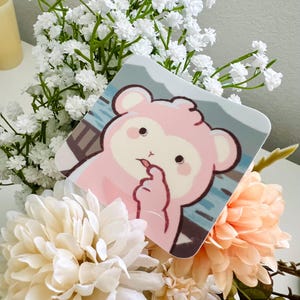 May include: A square sticker with rounded corners featuring a cartoon illustration of a pink bear-like creature with a cream-colored face, making a finger heart gesture against a background of blue water and a dark gray mountain silhouette.