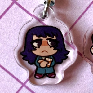 May include: A clear acrylic keychain featuring a cartoon character with purple hair, a teal shirt, and red shoes. The character is holding a white object in their hands.