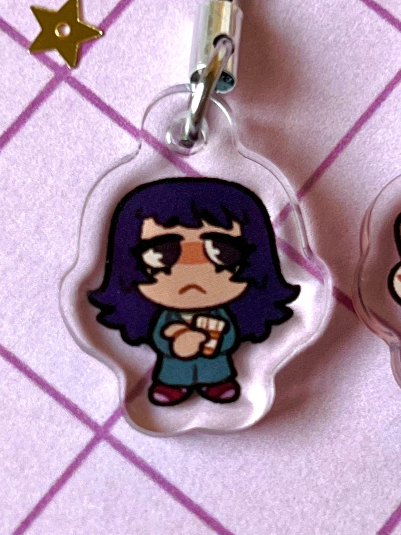 May include: A clear acrylic keychain featuring a cartoon character with purple hair, a teal shirt, and red shoes. The character is holding a white object in their hands.