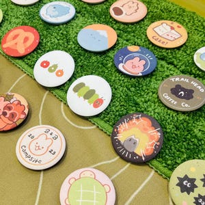 Può includere: Una collezione di spille rotonde e colorate con varie illustrazioni di cartoni animati. I disegni includono un pretzel, un bruco, un orso con caffè, un coniglietto in tuta spaziale e una torta. Le spille sono disposte su una superficie verde strutturata.