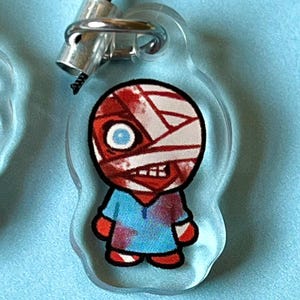 May include: A clear acrylic keychain with a cartoon character. The character is a person with a red and white bandage on their head, a blue shirt with red splatters, and red pants. The character has a blue eye and a red mouth.