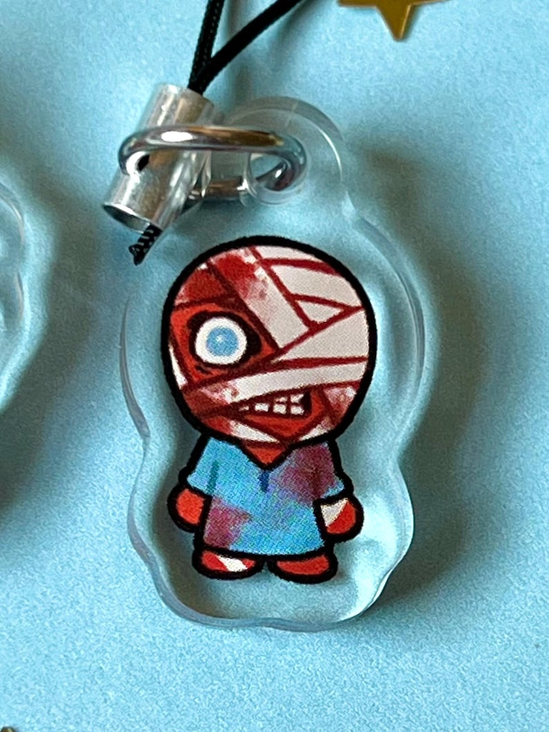 May include: A clear acrylic keychain with a cartoon character. The character is a person with a red and white bandage on their head, a blue shirt with red splatters, and red pants. The character has a blue eye and a red mouth.