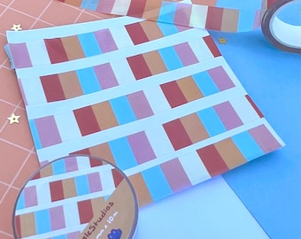 May include: A roll of decorative tape with a colorful rectangular pattern, alongside strips of the same tape and a sheet of paper with the same design. The tape roll is labeled "mkStudios 15mm x 10m". The background is a mix of pink, blue, and white.