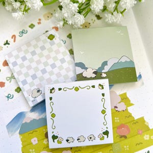 May include: Three square sticky note pads featuring sheep illustrations. One pad has a light blue and white checkerboard pattern with a sheep, one shows a green landscape with mountains and sheep, and one has a white center with a green floral border.