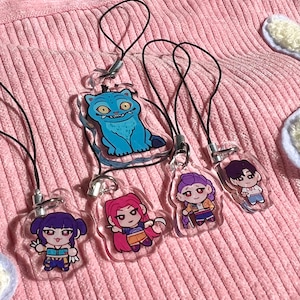 May include: A collection of clear acrylic charms featuring cartoon characters and a blue monster design. Each charm has a black cord for attachment. The charms are displayed on a pink, textured surface.