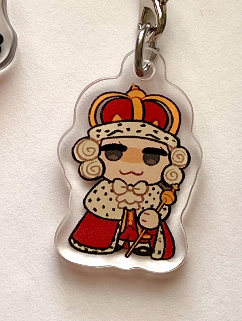 K&ouml;nnte beinhalten: Ein durchsichtiger Acryl-Schl&uuml;sselanh&auml;nger mit einer Comicfigur, die eine rot-goldene Krone, eine wei&szlig;-goldene Robe tr&auml;gt und ein goldenes Zepter h&auml;lt.