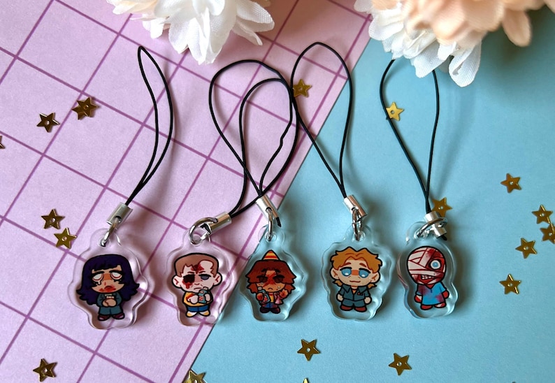 May include: Five colorful character phone charms with black cords. The charms are made of clear plastic with cartoon characters inside. The characters are a girl with black hair, a boy with brown hair, a boy with orange hair, a boy with blonde hair, and a boy with bandages on his head.