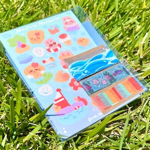 Könnte beinhalten: Eine transparente Kunststoffverpackung mit Aufklebern und dekorativem Klebeband zum Thema Nautik. Die Aufkleber zeigen Zeichentricktiere, Eiscreme und ein Boot. Das Klebeband hat Wellen-, Fisch- und Streifendesigns. Der Text "Nautical Nonsense" ist sichtbar.