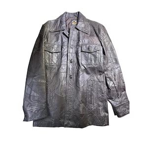 May include: A dark brown leather jacket with a patchwork design. The jacket has a collared neckline, two chest pockets, and two lower pockets. It features button closures and long sleeves. The jacket's texture appears worn, giving it a vintage aesthetic.