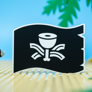 May include: A black pirate flag with a white skull and crossbones design. The skull is replaced with a white cup with a stem and a bone at the bottom. The flag is on a sandy surface with a Lego pirate figure in the foreground.