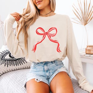 May include: Cream-colored long-sleeve shirt featuring a large red bow design. The bow has a watercolor effect with white accents. The shirt is paired with denim shorts. The overall aesthetic is casual and comfortable.