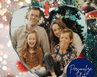 May include: Heart-shaped crystal ornament with a family photo inside. The ornament has a red ribbon for hanging. A dark blue circle with the words "Personalize Photo" is at the bottom right. The background has bokeh lights and a Christmas tree.