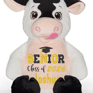 May include: A plush cow toy with a white body, black patches, and black hooves. The cow has embroidered text that reads "SENIOR Class of 2026 Joshua" in gold and black, with a graduation cap graphic. The cow has a pink tongue sticking out.