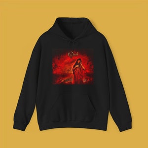 May include: Black hoodie with a red and gold graphic of a woman in a red dress standing in a forest with a cross in the background. The text "Opeth" is printed in gold above the image.