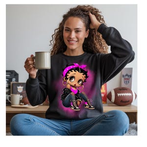 Puede incluir: Sudadera negra de cuello redondo con una ilustración de dibujos animados de una mujer con una diadema rosa y aros. El gráfico está sobre un fondo rosa. La mujer lleva un top rosa, una chaqueta negra y vaqueros rotos.