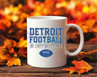 Könnte beinhalten: Weiße Keramiktasse mit dem Text "DETROIT FOOTBALL" in blauem Glitzer-Schriftzug und einer blauen Glitzer-Football-Grafik. Der Satz "IN GRIT WE TRUST" steht darunter. Die Tasse steht auf einer Holzoberfläche mit Herbstblättern.