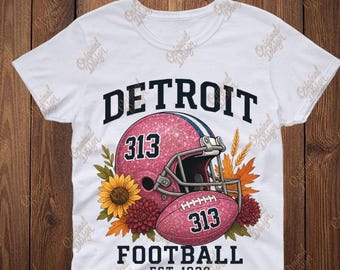 Könnte beinhalten: Weißes T-Shirt mit einem pinkfarbenen Glitzer-Footballhelm und -ball, beide mit der Nummer 313. Das Wort "DETROIT" steht über dem Helm und "FOOTBALL EST. 1930" darunter. Blumige Akzente umgeben Helm und Ball.