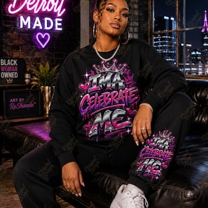 Può includere: Un completo composto da felpa a maniche lunghe e pantaloni della tuta neri con una stampa grafica che recita "I'MA CELEBRATE ME" in lettere graffiti argento metallizzato e viola, arricchito da cuori rosa, stelle e un disegno a corona.