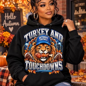 May include: A black hooded sweatshirt featuring a graphic of a lion wearing a blue Detroit football cap, holding a turkey leg in its mouth. The text reads TURKEY AND TOUCHDOWNS, with footballs and autumn leaves surrounding the central lion illustration.