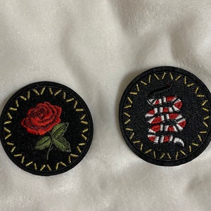 May include: Two black embroidered iron-on patches. One patch features a red rose with green leaves. The other patch features a black and white snake with red stripes.