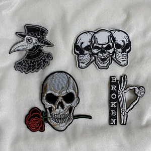 May include: Embroidered patches featuring gothic and macabre designs. Includes a plague doctor, three skulls, a skull with a rose, a skeleton hand with a heart, and the word "BROKEN". The patches are black, white, and red.