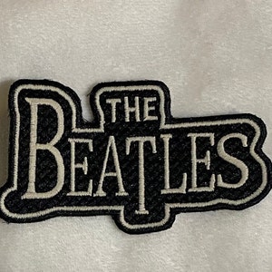 May include: A black and white embroidered patch featuring the text "THE BEATLES". The patch has a black background with white lettering and a white border. The patch is rectangular with rounded edges.