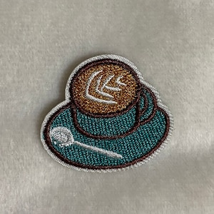 May include: A brown and teal embroidered patch of a coffee cup with a white spoon and a brown leaf design.