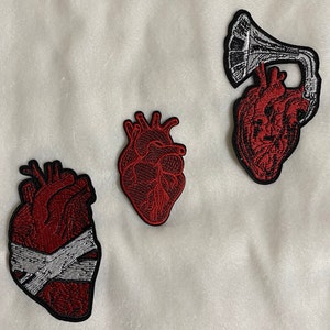 May include: Three embroidered iron-on patches featuring anatomical hearts. One heart is red with a white bandage, another is red with a detailed design, and the third is red with a white musical instrument.