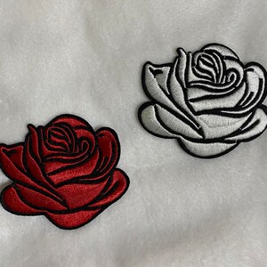 May include: Two embroidered rose patches. One is a deep red with black outlining, and the other is white with black outlining. Both patches have a detailed floral design and are set against a white background.