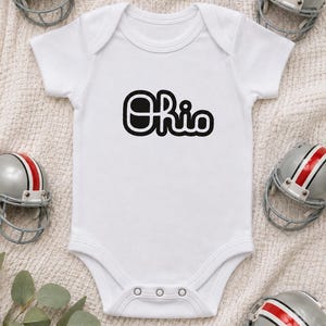 May include: A white short-sleeved baby bodysuit featuring the word Ohio printed in a bold black rounded font across the chest. The garment has a crew neckline and three metal snap closures at the bottom for easy access and secure fastening.