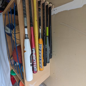 Może przedstawiać: Drewniany organizer na sprzęt sportowy zamontowany na ścianie, mieszczący kilka kij&oacute;w baseballowych i piłek sportowych. Kije są w r&oacute;żnych kolorach, w tym białym, czerwonym, ż&oacute;łtym i czarnym. Organizer zawiera r&oacute;wnież koszyk&oacute;wki i inny sprzęt sportowy.