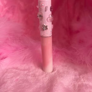 May include: A pink lip gloss tube with a white and pink gradient. The top is decorated with various embellishments, including rhinestones, cat-shaped charms, and flower-shaped charms. The background is a soft, pink, feathery texture.