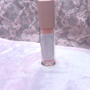 May include: A clear lip gloss tube with a light pink tint and a matching pink cap. The label has text and is set against a soft, white background. The background is a white, textured fabric.