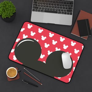 May include: A rectangular desk mat featuring a vibrant red background covered in a repeating pattern of small white Mickey Mouse head silhouettes, with a large black Mickey Mouse head shape prominently displayed in the center.
