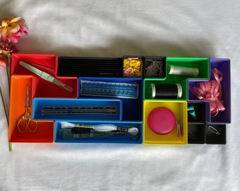 May include: A set of colorful plastic storage containers with various sewing supplies, including thread, needles, scissors, and a measuring tape. The containers are arranged in a grid pattern on a white surface.