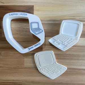 May include: A white plastic cookie cutter shaped like a laptop computer. The cutter has a small handle on the side and the text "Laptop | A2EH" is printed on the top. There is a smaller white plastic cookie cutter shaped like a laptop computer on a wooden surface.